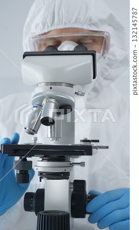 Male scientist virologist wearing hazmat suit, mask, and blue gloves, diligently working at a microscope in a laboratory researching viruses. Medicine and science concept Male scientist virologist wearing hazmat suit, mask, and blue gloves, diligently working at a microscope in a laboratory researching viruses. Medicine and science concept 132145787