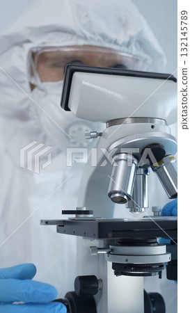 Male scientist virologist wearing hazmat suit, mask, and blue gloves, diligently working at a microscope in a laboratory researching viruses. Medicine and science concept Male scientist virologist wearing hazmat suit, mask, and blue gloves, diligently working at a microscope in a laboratory researching viruses. Medicine and science concept 132145789