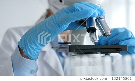 Close up of scientist's gloved hands adjusting microscope, conducting research in laboratory with test tubes containing blue liquid in background. Medicine, healthcare and science concept Close up of scientist's gloved hands adjusting microscope, conducting research in laboratory with test tubes containing blue liquid in background. Medicine, healthcare and science concept 132145801