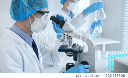 Masked scientist examining specimen under microscope while laboratory assistant handles glassware in a professional medical investigation. Health care and science concept 132145808