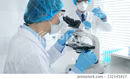 Woman scientist in blue protective gear is examining specimen under microscope while laboratory assistant handles glassware in medical investigation at the background. Medicine, and science concept Woman scientist in blue protective gear is examining specimen under microscope while laboratory assistant handles glassware in medical investigation at the background. Medicine, and science concept 132145817