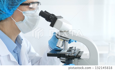 Female scientist analyzing research sample through microscope, wearing protective lab gear for medical investigation. Laboratory and science concept 132145818