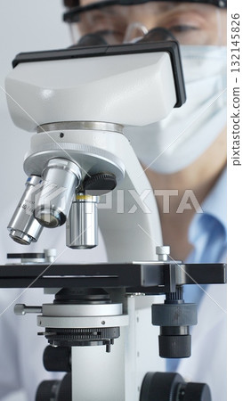 Scientist wearing lab coat and protective glasses using a modern microscope in a laboratory setting, conducting research and experiments in a scientific environment. Health care and science concept 132145826