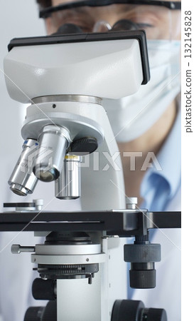Scientist wearing lab coat and protective glasses using a modern microscope in a laboratory setting, conducting research and experiments in a scientific environment. Health care and science concept 132145828