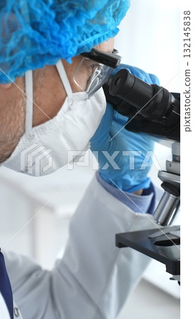 Medical scientist senior man wearing protective mask, glasses and bouffant cap using microscope doing scientific research in a laboratory. Medicine and science concept Medical scientist senior man wearing protective mask, glasses and bouffant cap using microscope doing scientific research in a laboratory. Medicine and science concept 132145838