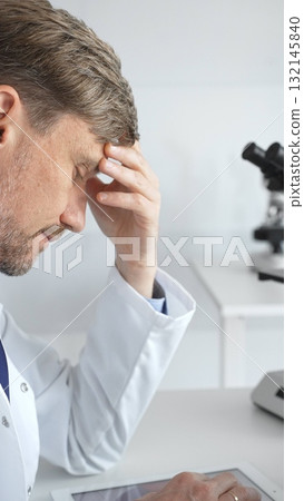 Confident male mature scientist is thinking and analyzing data on a tablet in his laboratory, with a microscope in the background, suggesting advanced scientific research. Medicine and science concept 132145840