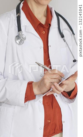 Close up of woman doctor wearing stethoscope and white coat over brick red blouse is standing and writing on a clipboard, representing medical expertise and patient care. Medicine concept 132145841