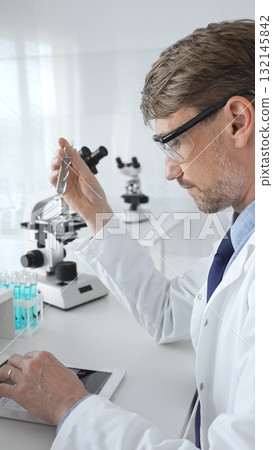 Male researcher in protective glasses holding test tube with blue liquid and working with tablet in sterile laboratory environment. Medicine and science concept 132145842