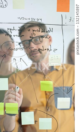 Business colleagues brainstorming, writing strategy notes on glass board, collaborating with colorful sticky notes in creative workplace meeting Business colleagues brainstorming, writing strategy notes on glass board, collaborating with colorful sticky notes in creative workplace meeting 132145897