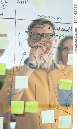 Business colleagues brainstorming, writing strategy notes on glass board, collaborating with colorful sticky notes in creative workplace meeting Business colleagues brainstorming, writing strategy notes on glass board, collaborating with colorful sticky notes in creative workplace meeting 132145918