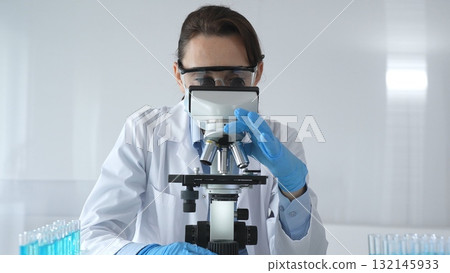 A female scientist in a lab coat and blue gloves examines a microscope slide in a sterile laboratory, conducting research using blue samples from a test tube 132145933