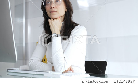 Businesswoman wearing white casual sweater and glasses is sitting and thinking near computer monitor screen in a modern, clean office environment. Audit in business 132145957