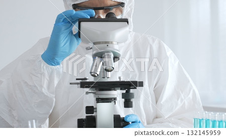 Male doctor in protective suit and gloves using microscope while analyzing samples while conducting research in sterile laboratory. Close-up of test tubes with blue liquid Male doctor in protective suit and gloves using microscope while analyzing samples while conducting research in sterile laboratory. Close-up of test tubes with blue liquid 132145959