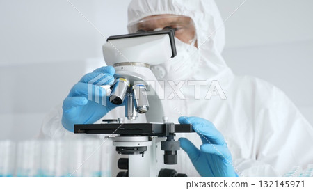 Male scientific researcher or virologist wearing protective gear adjusting microscope, analyzing lab samples with precision in sterile research environment. Medicine concept Male scientific researcher or virologist wearing protective gear adjusting microscope, analyzing lab samples with precision in sterile research environment. Medicine concept 132145971