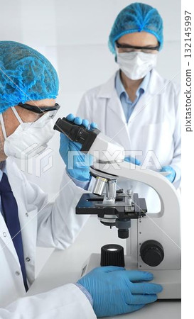 Two scientists in blue protective hairnets, masks and glasses conduct research under a microscope in a laboratory. Concept of medicine and science 132145997