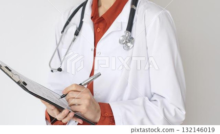Unknown woman doctor wearing stethoscope and white coat over brick red blouse is standing and writing on a clipboard, representing medical expertise and patient care. Medicine and health care concept Unknown woman doctor wearing stethoscope and white coat over brick red blouse is standing and writing on a clipboard, representing medical expertise and patient care. Medicine and health care concept 132146019