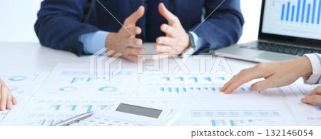 Business people reviewing financial charts and reports in office, using a laptop and calculator while engaging in discussions about strategy and performance 132146054
