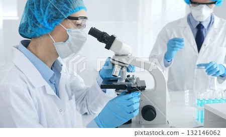 Woman scientist in blue protective gear is examining specimen under microscope while laboratory assistant handles glassware in medical investigation at the background. Medicine, and science concept Woman scientist in blue protective gear is examining specimen under microscope while laboratory assistant handles glassware in medical investigation at the background. Medicine, and science concept 132146062