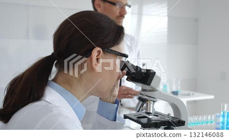 A female scientist wearing protective glasses looks into a microscope and conducts a medical research in a sterile laboratory while her male colleague works in the background. Medicine and science 132146088