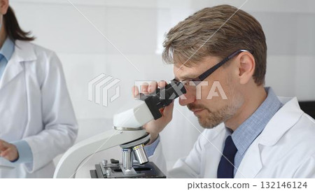 A researcher wearing glasses examines a sample under a microscope, a lab technician wearing a protective mask stands nearby, both in white lab coats during the study. Medicine and Science 132146124