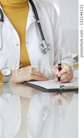 Doctor wearing stethoscope and yellow sweater under white lab coat, making notes on a clipboard in a bright medical office. Medicine, healthcare and science concept Doctor wearing stethoscope and yellow sweater under white lab coat, making notes on a clipboard in a bright medical office. Medicine, healthcare and science concept 132146131