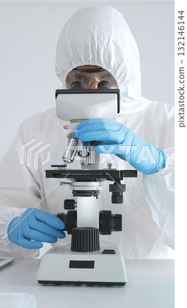 Medical scientist in protective suit using microscope while analyzing samples, conducting research in sterile laboratory environment. Close-up of test tubes with blue liquid 132146144