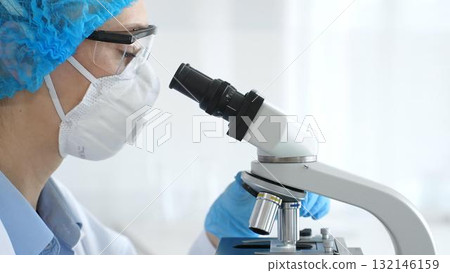 Woman researcher in blue colored protective gear examining sample through microscope, conducting medical experiment inside laboratory with glass vials surrounding workstation. Medicine and science Woman researcher in blue colored protective gear examining sample through microscope, conducting medical experiment inside laboratory with glass vials surrounding workstation. Medicine and science 132146159