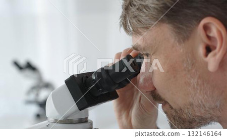 Close up of a man scientist carefully observing through a microscope in a brightly lit laboratory, conducting research for developing new medicines and treatments. Medicine and science concept 132146164