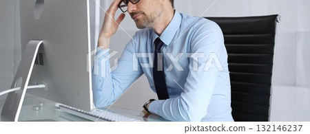Overworked businessman experiencing headache and painful migraine, feeling overwhelming stress and burnout while working long hours at computer in a modern office setup 132146237