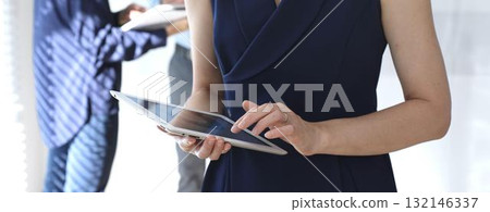 Professional businesswoman using a digital tablet in a modern office, interacting with a touchscreen while blurred colleagues collaborate in the background, focusing on tech and teamwork 132146337