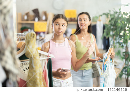 Stubborn teen girl is moody and shows dissatisfaction while shopping in cloth store with her mother Stubborn teen girl is moody and shows dissatisfaction while shopping in cloth store with her mother 132146526