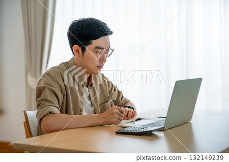 Happy young Asian man working on laptop in living room Happy young Asian man working on laptop in living room 132149239