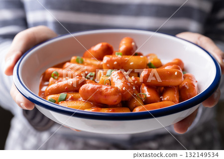 Korean Tteokbokki (simmered rice cake) and sausage with spicy sauce in bowl holding by hand Korean Tteokbokki (simmered rice cake) and sausage with spicy sauce in bowl holding by hand 132149534