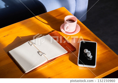 Coffee cup, open notebook, phone and earbud on wood table. 132150960
