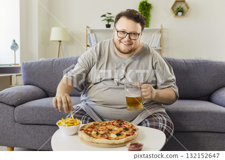 Overweight man at home eating pizza and fries with beer on living room sofa 132152647