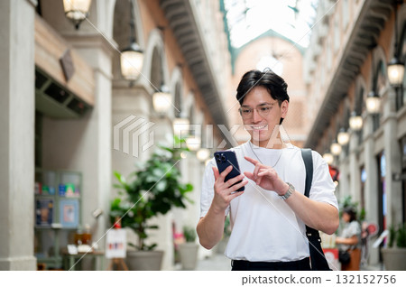 Asian man with glasses in white tee is smiling while looking at his phone in shopping mall or market 132152756