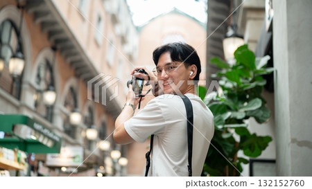 Asian man wearing glasses looking back while taking a photo in hallway of shopping mall or market. 132152760