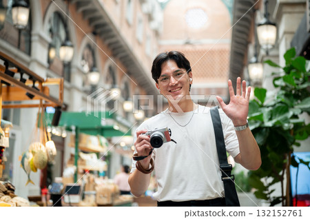 Asian man wearing glasses waving a hand while holding a camera in hallway of shopping mall or market 132152761