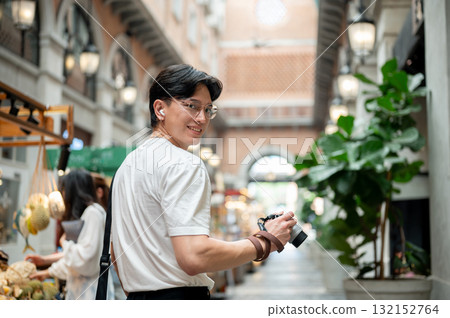 Asian man wearing glasses is looking back while holding camera in hallway of shopping mall or market Asian man wearing glasses is looking back while holding camera in hallway of shopping mall or market 132152764