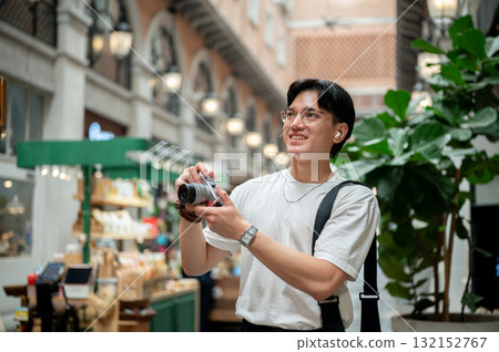 Asian man wearing glasses in white tee looking around with camera in shopping mall or indoor market. Asian man wearing glasses in white tee looking around with camera in shopping mall or indoor market. 132152767
