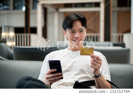 Asian man wearing glasses in white tee holding phone and looking at credit card while sit on sofa. Asian man wearing glasses in white tee holding phone and looking at credit card while sit on sofa. 132152784
