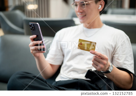 Asian man wearing glasses in white t shirt holding smartphone and credit card while sitting in cafe. 132152785