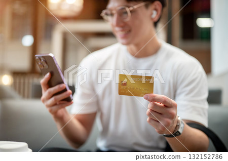 Asian man wearing glasses in white shirt is holding smartphone and credit card while sitting in cafe 132152786
