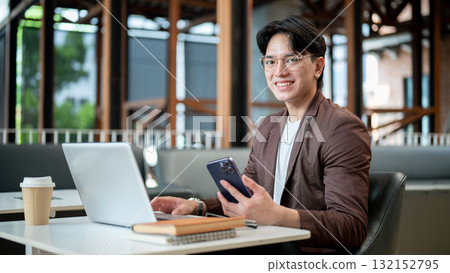 Brown suit asian man with glasses is smiling while holding his phone sitting with laptop in the cafe Brown suit asian man with glasses is smiling while holding his phone sitting with laptop in the cafe 132152795