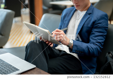 Blue suit asian man is writing with a pen in notebook while sitting cross legged on sofa in the cafe 132152812