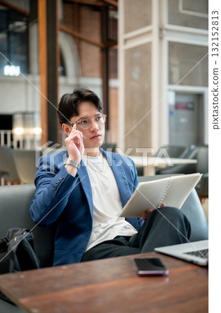 Blue suit asian man holding notebook and thinking thoughtfully while sitting at wooden table in cafe 132152813