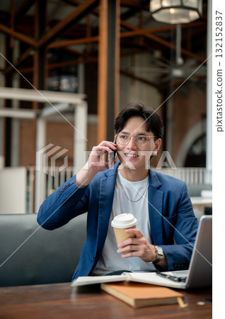 Blue suit man talking on phone while holding coffee cup, sitting with laptop in cafe or coffee shop. 132152837