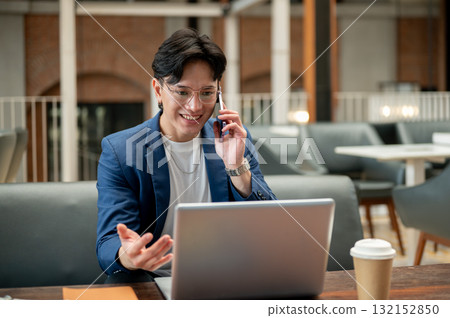 Asian man in blue suit is talking on the phone while sitting with a laptop in a cafe or coffee shop. 132152850