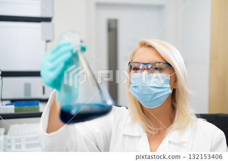 Blonde woman scientist in laboratory wearing white lab coat carefully examining blue solution in glass flask. Blonde woman scientist in laboratory wearing white lab coat carefully examining blue solution in glass flask. 132153406