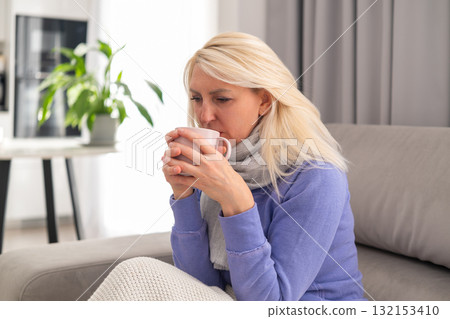 Middle aged blonde woman with scarf around her neck, sitting on sofa and holding cup of tea. Concept of flu, cold, home rest, and recovery. 132153410